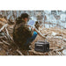 Person in camouflage jacket using a tablet in a forest setting with an Aeon Guardians G1200 portable power station nearby.